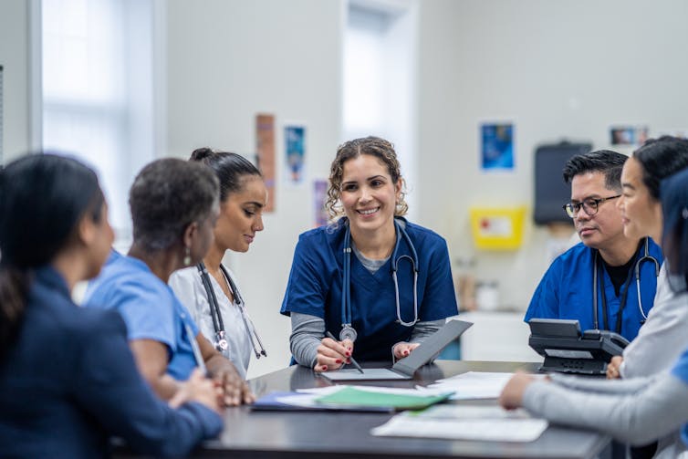 A small group of medical professionals gather around a table. They are each dressed professionally and have files scattered between them.