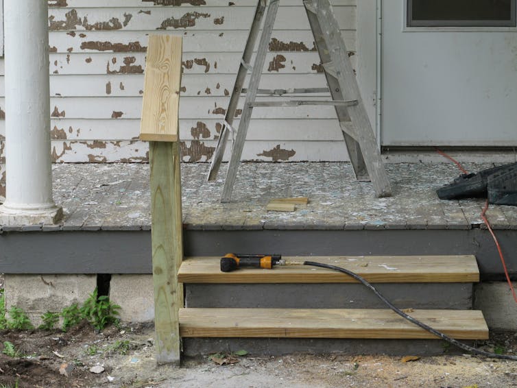 An older home with pealing paint on the exterior walls behind a porch. Construction equipment sits on a new step being built to the porch.