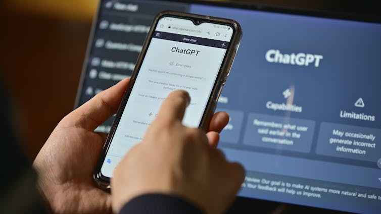 A person asks ChatGPT questions on a phone screen while a laptop open to the ChatGPT landing page is open in the background